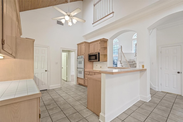 kitchen featuring ceiling fan, a towering ceiling, light brown cabinetry, tile counters, and kitchen peninsula