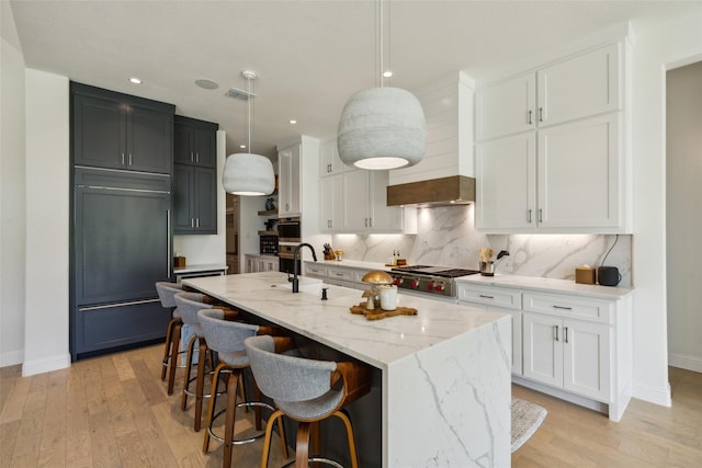 kitchen featuring a kitchen island with sink, white cabinets, hanging light fixtures, light stone countertops, and appliances with stainless steel finishes