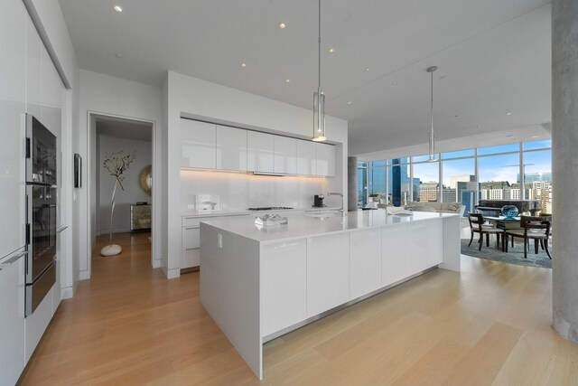 kitchen with pendant lighting, a center island with sink, oven, white cabinets, and light wood-type flooring