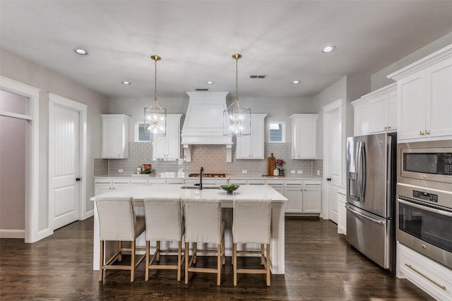 kitchen featuring premium range hood, hanging light fixtures, stainless steel appliances, white cabinets, and a center island with sink