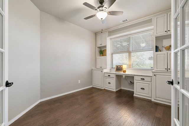 unfurnished office featuring visible vents, a ceiling fan, dark wood finished floors, built in desk, and baseboards