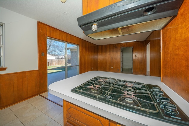 kitchen featuring a textured ceiling, wooden walls, exhaust hood, light tile patterned floors, and stainless steel gas stovetop