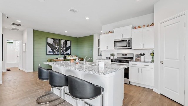 kitchen with light stone countertops, stainless steel appliances, a kitchen island with sink, light hardwood / wood-style floors, and white cabinetry