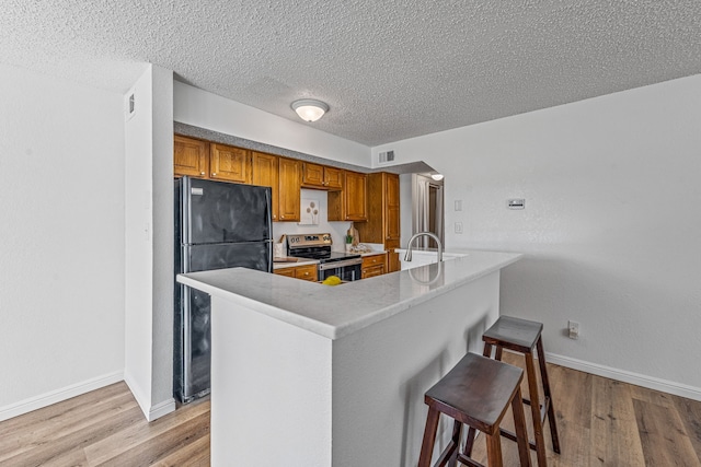 kitchen featuring a breakfast bar area, brown cabinetry, stainless steel electric range, freestanding refrigerator, and light wood-style floors