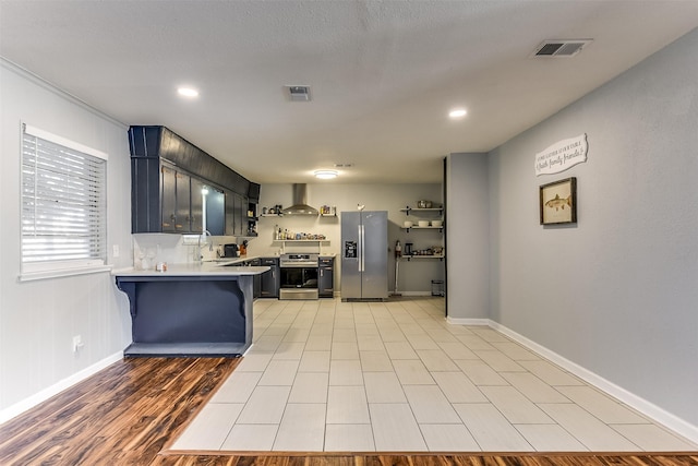 kitchen with kitchen peninsula, appliances with stainless steel finishes, a breakfast bar, blue cabinets, and wall chimney range hood