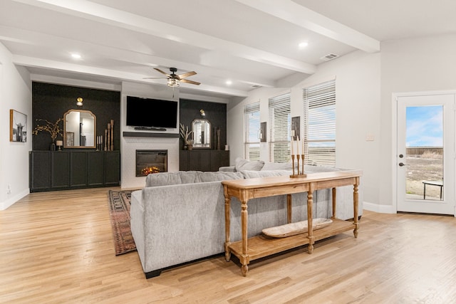 living area featuring a glass covered fireplace, beam ceiling, light wood-style flooring, and baseboards