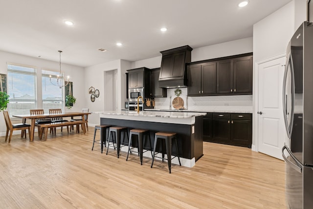 kitchen featuring premium range hood, appliances with stainless steel finishes, hanging light fixtures, a kitchen island with sink, and light hardwood / wood-style flooring