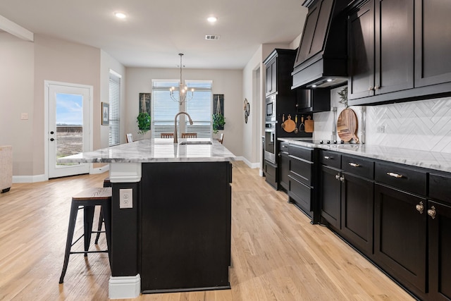 kitchen featuring a kitchen breakfast bar, light stone countertops, decorative backsplash, a center island with sink, and decorative light fixtures