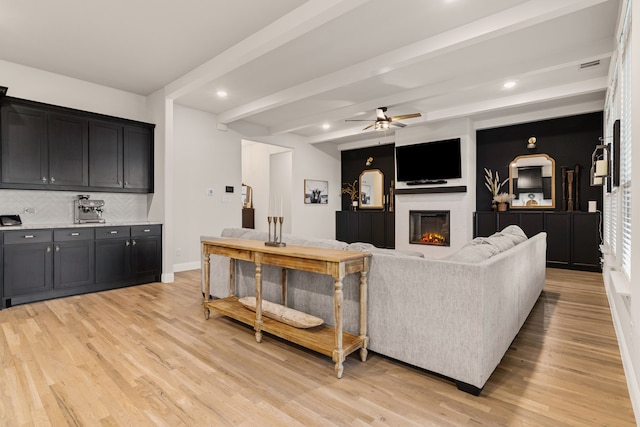 living room with ceiling fan, beamed ceiling, a glass covered fireplace, and light wood-style flooring