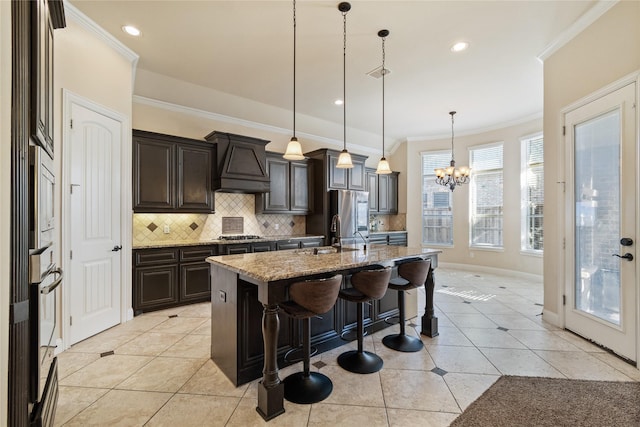 kitchen featuring hanging light fixtures, premium range hood, backsplash, a kitchen island with sink, and a breakfast bar