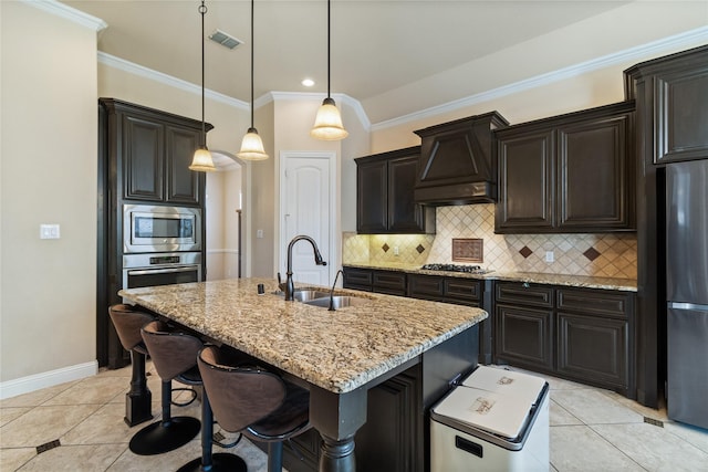 kitchen featuring premium range hood, a kitchen island with sink, hanging light fixtures, sink, and stainless steel appliances