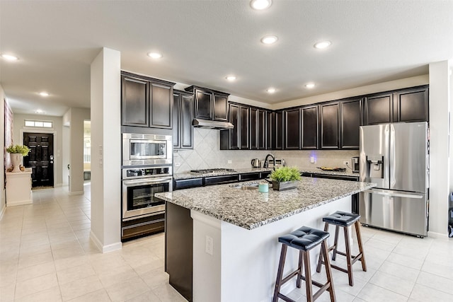 kitchen with light stone countertops, stainless steel appliances, sink, light tile patterned floors, and a center island with sink