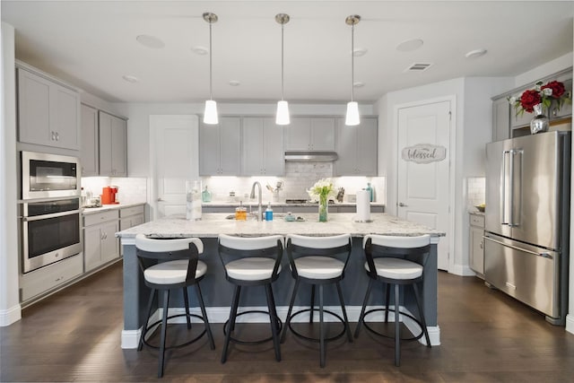 kitchen featuring appliances with stainless steel finishes, light stone counters, a kitchen island with sink, and pendant lighting