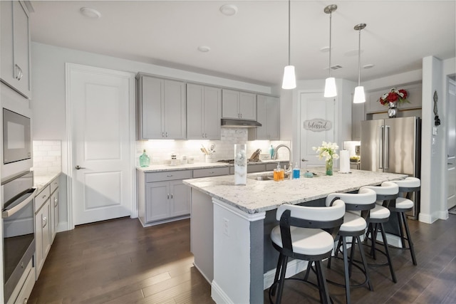 kitchen with a kitchen island with sink, light stone countertops, appliances with stainless steel finishes, decorative light fixtures, and dark hardwood / wood-style flooring