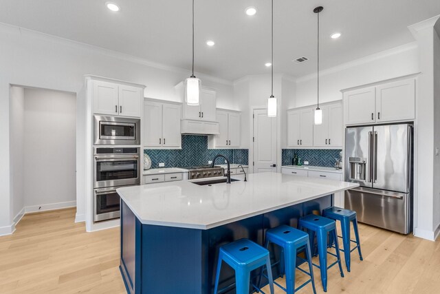 kitchen with appliances with stainless steel finishes, ornamental molding, white cabinetry, hanging light fixtures, and an island with sink