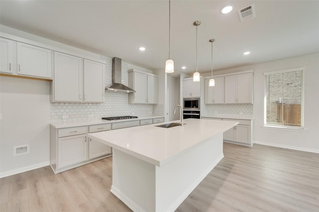 kitchen with appliances with stainless steel finishes, pendant lighting, sink, wall chimney range hood, and a center island with sink