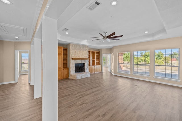 unfurnished living room featuring ceiling fan, a tray ceiling, crown molding, light wood-type flooring, and a stone fireplace