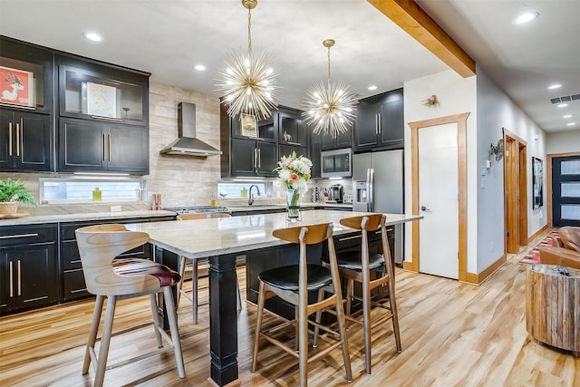 kitchen featuring appliances with stainless steel finishes, pendant lighting, an island with sink, light stone countertops, and wall chimney exhaust hood