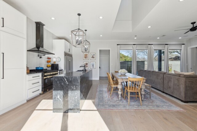 kitchen with light wood-type flooring, wall chimney exhaust hood, premium appliances, a kitchen island with sink, and white cabinetry