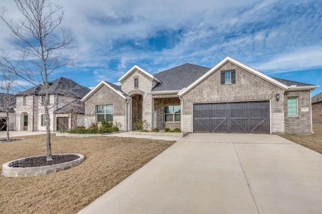 french country home with roof with shingles, an attached garage, concrete driveway, a front lawn, and brick siding