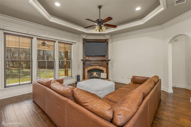 living room with dark wood-type flooring, a brick fireplace, ornamental molding, a tray ceiling, and ceiling fan