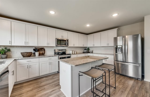 kitchen with light wood-type flooring, appliances with stainless steel finishes, a kitchen island, light stone countertops, and white cabinets