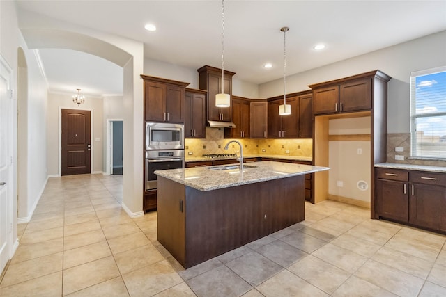 kitchen featuring appliances with stainless steel finishes, tasteful backsplash, light stone counters, hanging light fixtures, and an island with sink