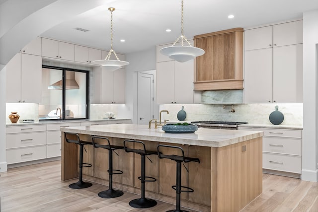 kitchen featuring pendant lighting, wall chimney range hood, white cabinetry, a kitchen island with sink, and tasteful backsplash