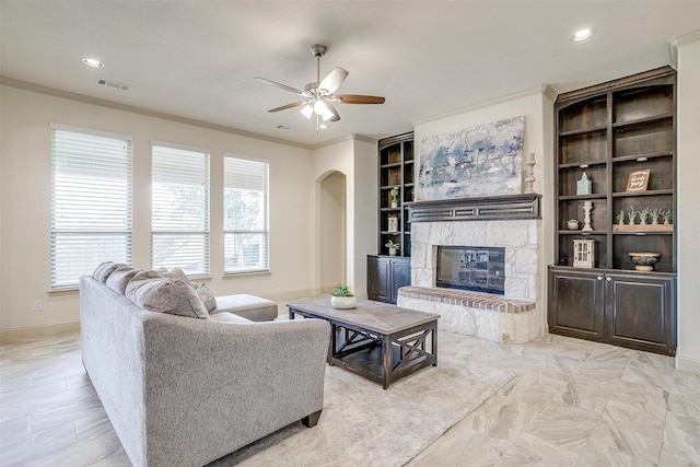 living room featuring ceiling fan, crown molding, a stone fireplace, and built in shelves