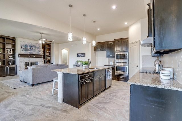 kitchen with sink, a kitchen island with sink, hanging light fixtures, stainless steel appliances, and light stone countertops