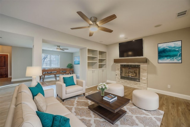 living room featuring built in shelves, ceiling fan, a tile fireplace, and light wood-type flooring