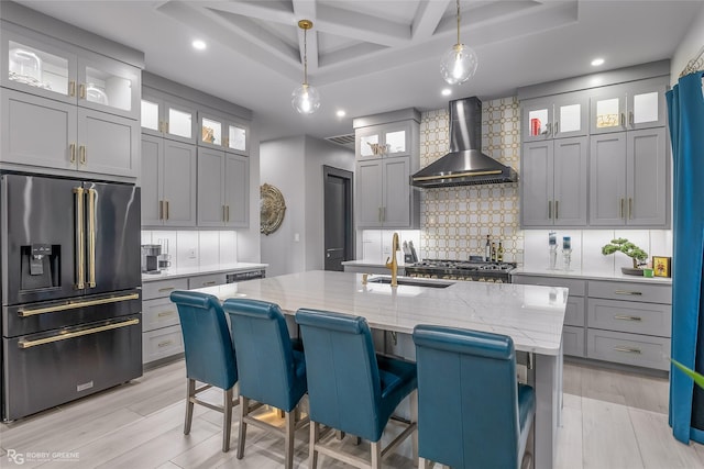 kitchen with pendant lighting, an island with sink, high end black fridge, light stone countertops, and wall chimney range hood