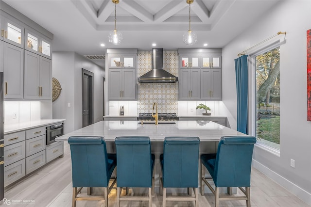 kitchen with backsplash, wall chimney range hood, a kitchen island with sink, and coffered ceiling