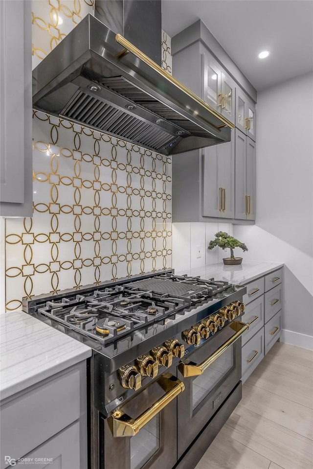 kitchen featuring decorative backsplash, light stone counters, gray cabinetry, wall chimney range hood, and double oven range