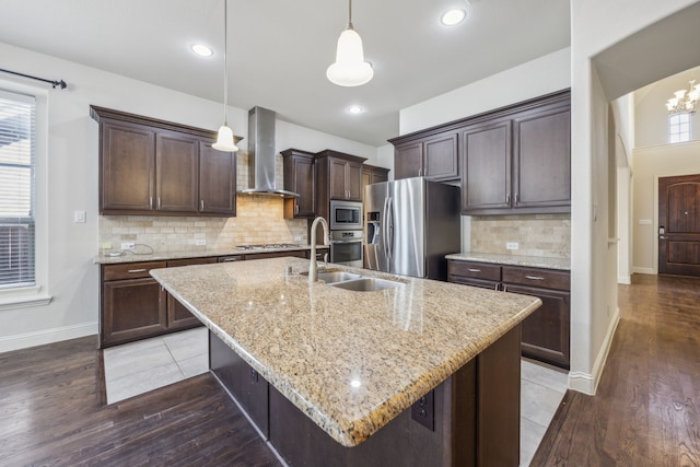 kitchen with appliances with stainless steel finishes, decorative light fixtures, an island with sink, dark brown cabinetry, and wall chimney range hood