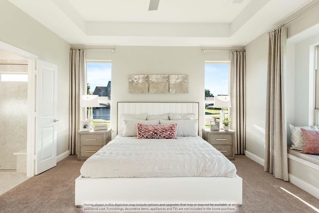 carpeted bedroom with a raised ceiling, multiple windows, ceiling fan, and ensuite bathroom