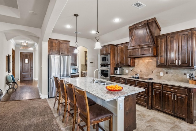 kitchen with arched walkways, decorative light fixtures, stainless steel appliances, premium range hood, and a center island with sink