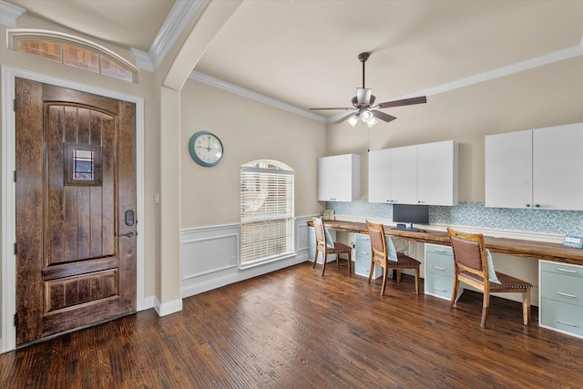 unfurnished office featuring ceiling fan, dark wood-style flooring, ornamental molding, wainscoting, and built in study area