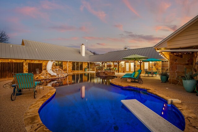 pool at dusk with a diving board, a patio area, and a water slide