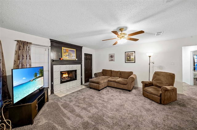 living room featuring a tiled fireplace, ceiling fan, light carpet, and a textured ceiling