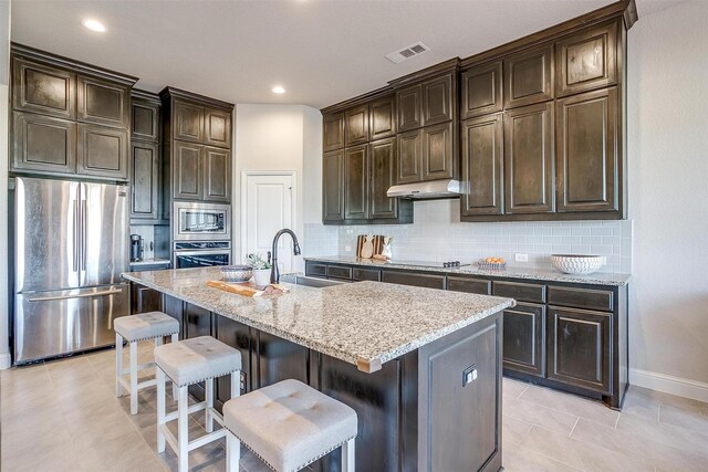 kitchen with dark brown cabinetry, sink, stainless steel appliances, an island with sink, and a kitchen bar