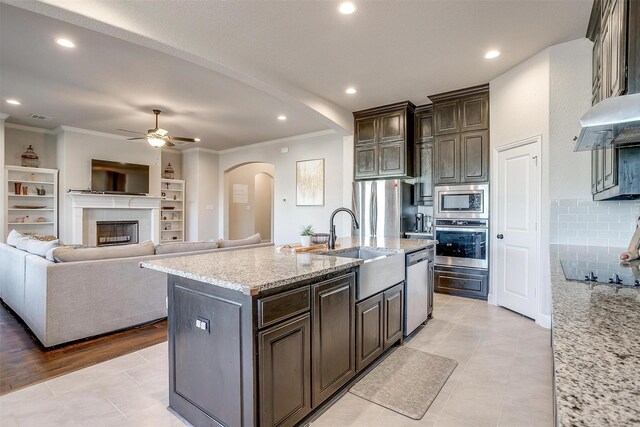 kitchen featuring light stone counters, dark brown cabinets, stainless steel appliances, a center island with sink, and light tile patterned flooring