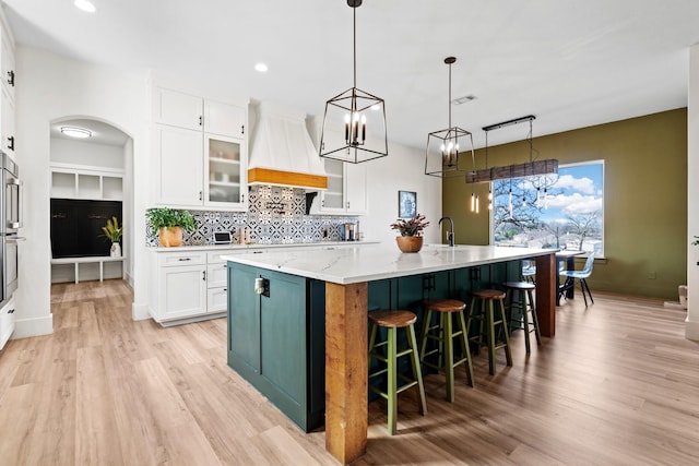 kitchen with light stone countertops, hanging light fixtures, a large island with sink, white cabinets, and custom range hood