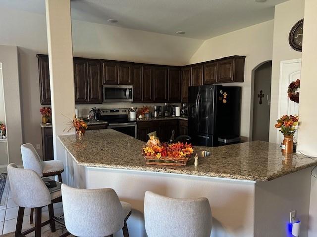 kitchen with dark brown cabinetry, light stone countertops, kitchen peninsula, a kitchen bar, and appliances with stainless steel finishes