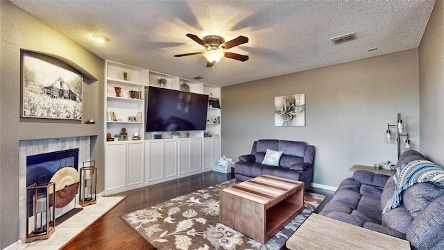 living room with ceiling fan, dark wood-type flooring, a tile fireplace, and a textured ceiling