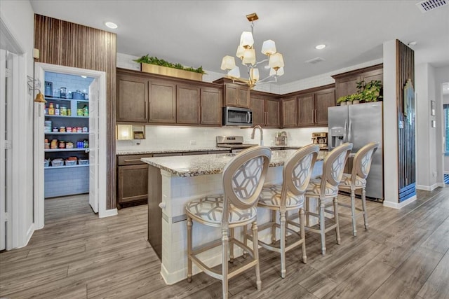 kitchen with light stone countertops, light hardwood / wood-style floors, pendant lighting, a center island with sink, and appliances with stainless steel finishes