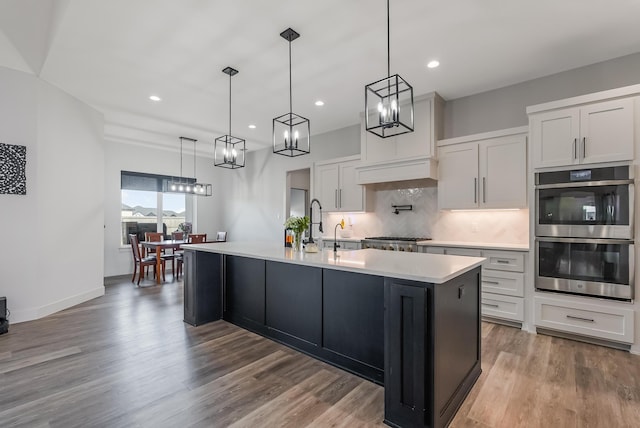 kitchen with a center island with sink, appliances with stainless steel finishes, decorative light fixtures, white cabinetry, and wood-type flooring