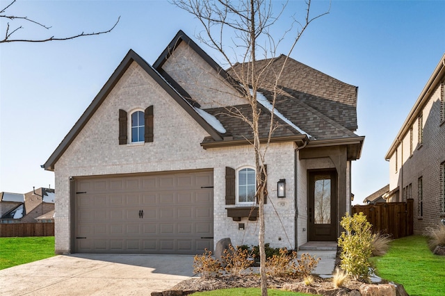 french country inspired facade with a garage, roof with shingles, concrete driveway, and fence