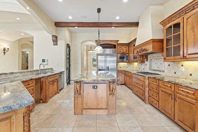 kitchen with light stone counters, custom exhaust hood, pendant lighting, built in appliances, and a center island with sink