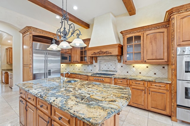 kitchen featuring beamed ceiling, custom range hood, built in appliances, and an island with sink